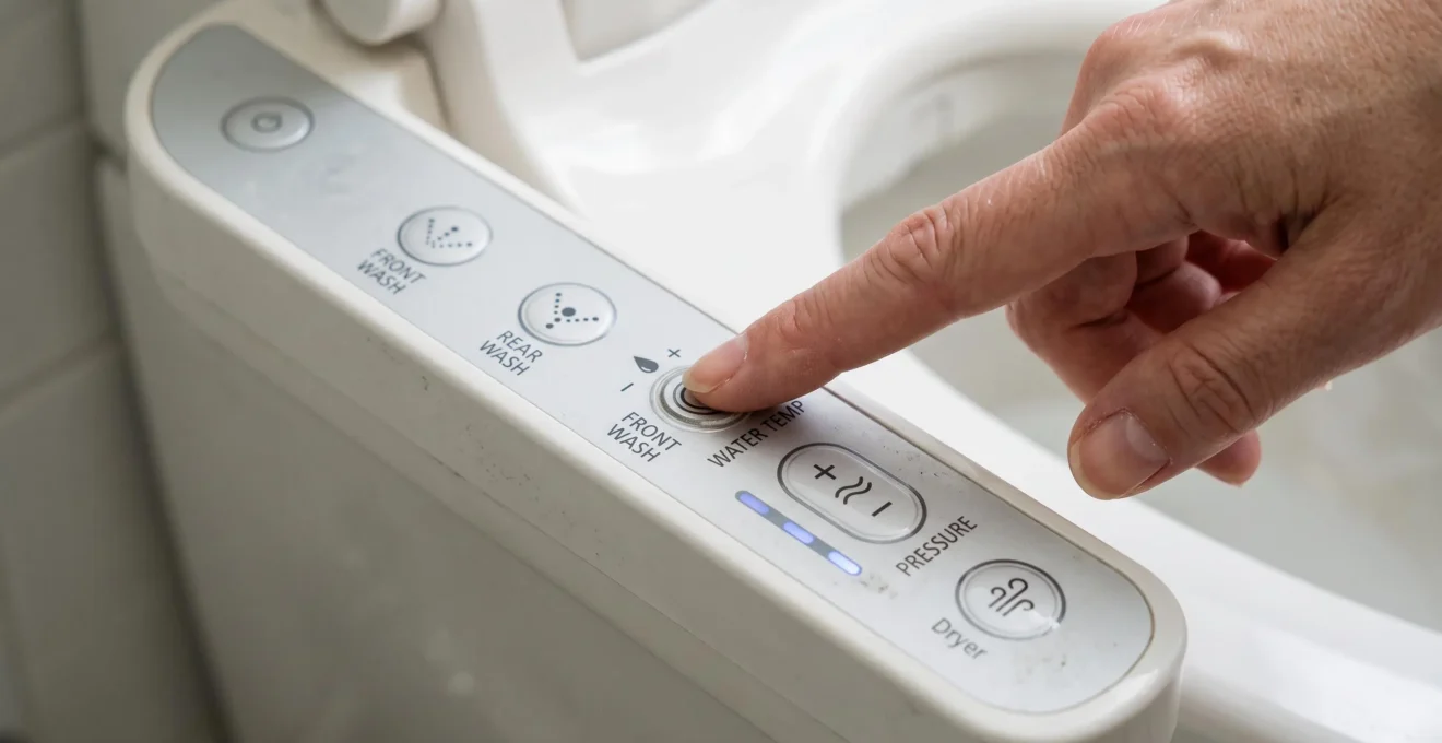 Gros plan sur une main appuyant sur le panneau de commande d'un WC japonais avec symboles de réglage température et jet d'eau